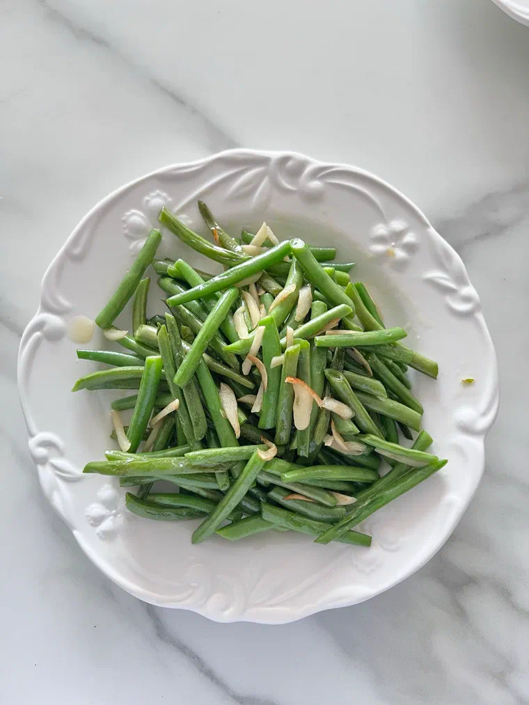 Une assiette blanche de haricots verts cuits parsemés d'amandes effilées, sur un plan en marbre