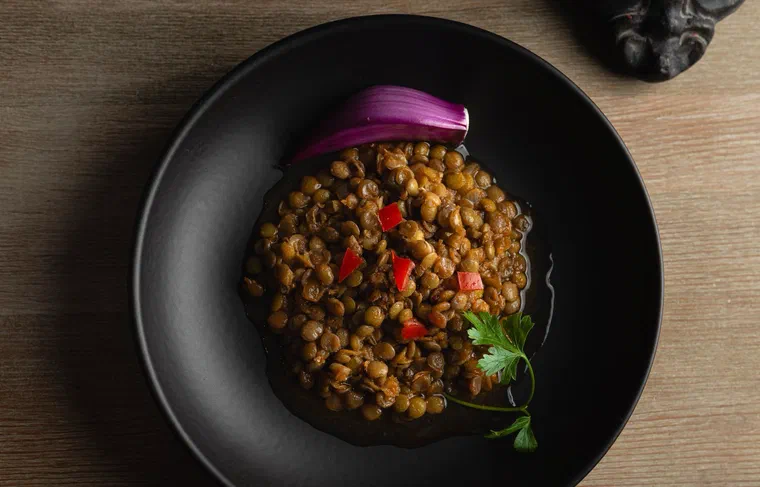 Lentilles vertes cuites dans une assiette creuse noire, garnies de dés de poivron rouge, de persil et d'un quartier d'oignon rouge