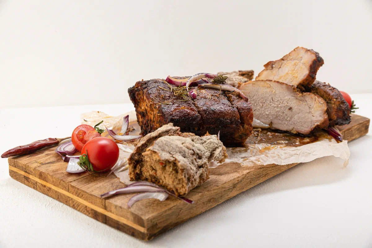 Un rôti de porc cuit à la croûte brune, en partie tranché, sur une planche en bois avec du pain, des tomates cerises et de l'oignon rouge