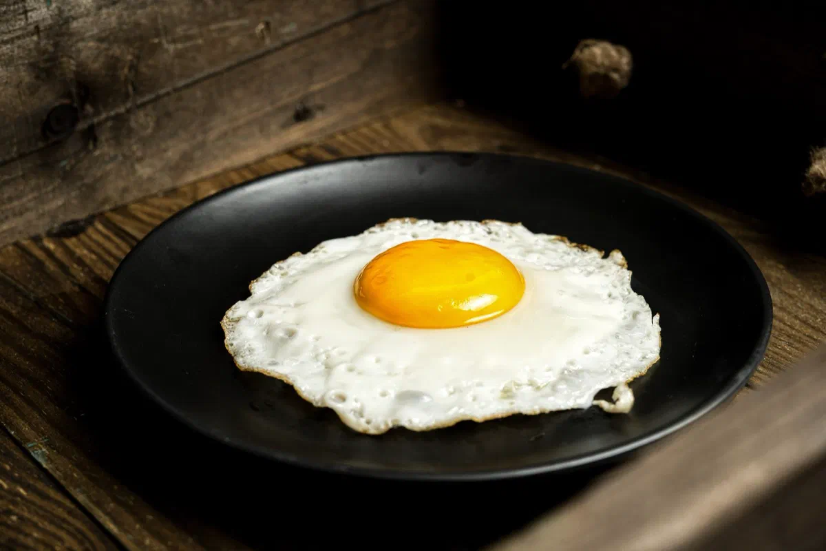 Un œuf au plat au blanc bien pris et au jaune bombé et brillant, seul dans une assiette noire posée sur une table en bois