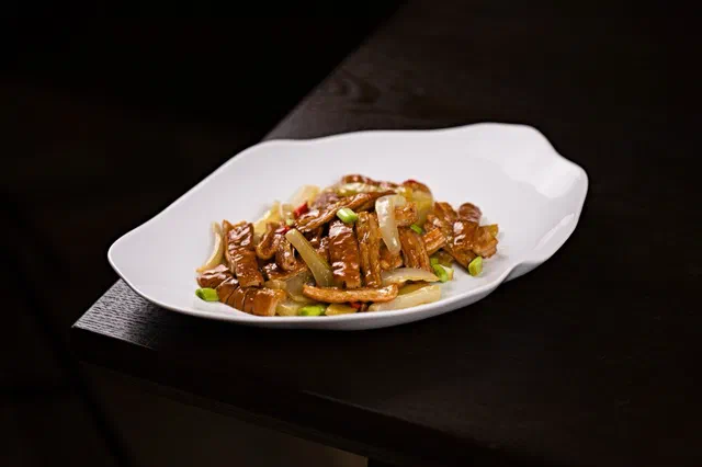 Lamelles de bœuf sautées avec des oignons émincés et quelques rondelles de ciboule, dans un plat blanc ondulé sur fond sombre