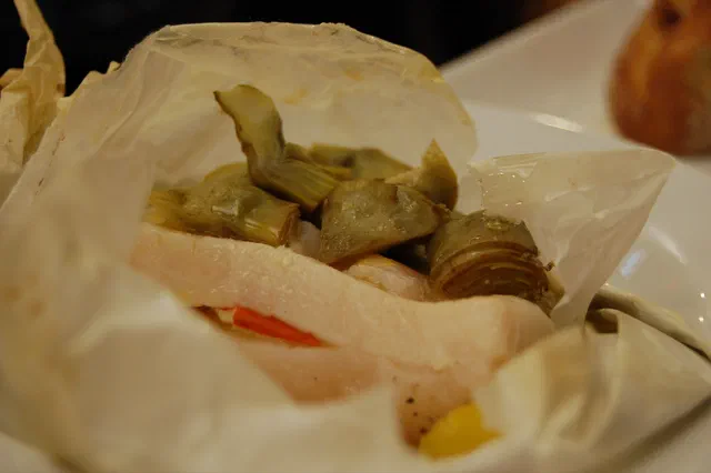 Papillote de papier cuisson ouverte sur un filet de cabillaud blanc, recouvert de cœurs d'artichaut et accompagné de lamelles de carotte
