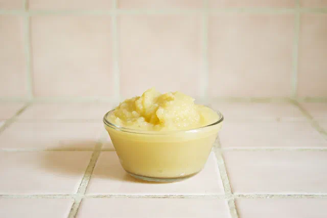Un petit bol en verre rempli de compote de pommes jaune pâle, posé sur un carrelage blanc