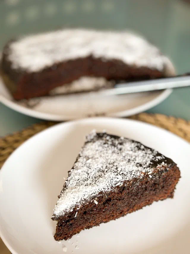 Une part de fondant au chocolat saupoudrée de sucre glace sur une assiette blanche, le reste du gâteau en arrière-plan