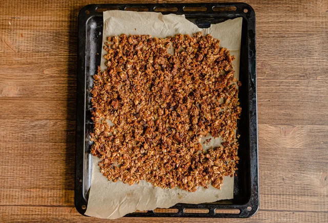 Granola maison doré en grappes étalé sur une plaque de four tapissée de papier cuisson, posée sur une table en bois