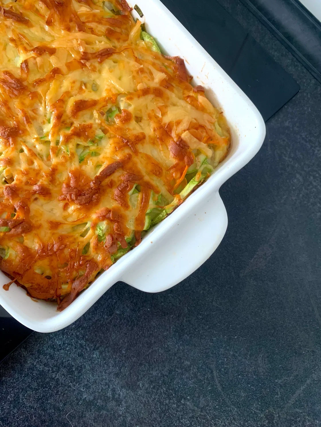 Un gratin de courgettes doré, gratiné au fromage, dans un plat à four blanc posé sur un plan de travail sombre