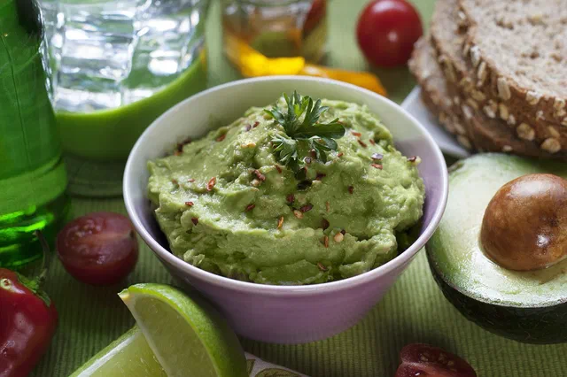 Un bol de guacamole parsemé de piment et d'une branche de persil, entouré d'un demi-avocat, de quartiers de citron vert et de tomates cerises