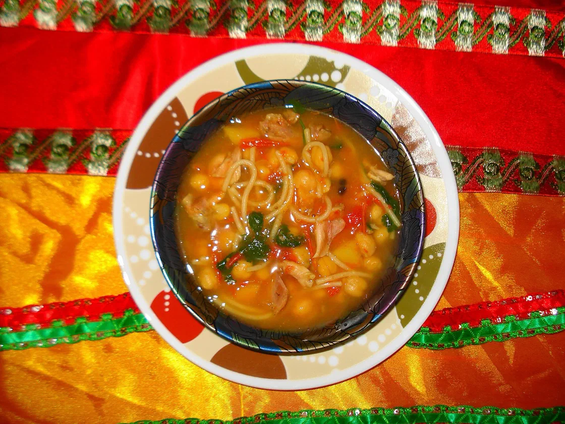 Un bol de harira vu de dessus, avec des pois chiches, des vermicelles, des morceaux de viande, de la tomate et des herbes dans un bouillon orangé, posé sur un tissu rouge et jaune