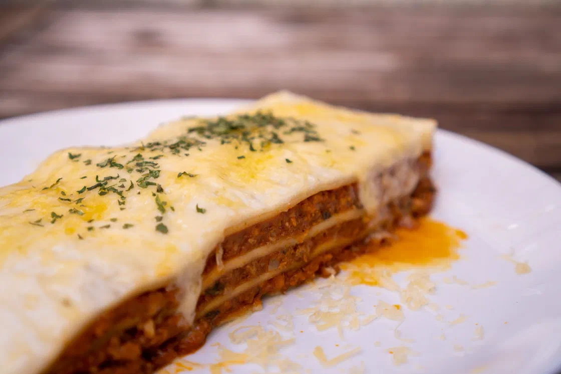Une part de lasagnes à la bolognaise sur une assiette blanche, nappée de fromage fondu et d'herbes, avec les couches de pâte et de viande visibles sur la tranche