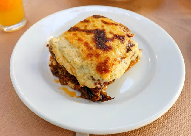 Une part de moussaka sur une assiette blanche, avec la béchamel dorée au-dessus et la couche de viande à la tomate en dessous