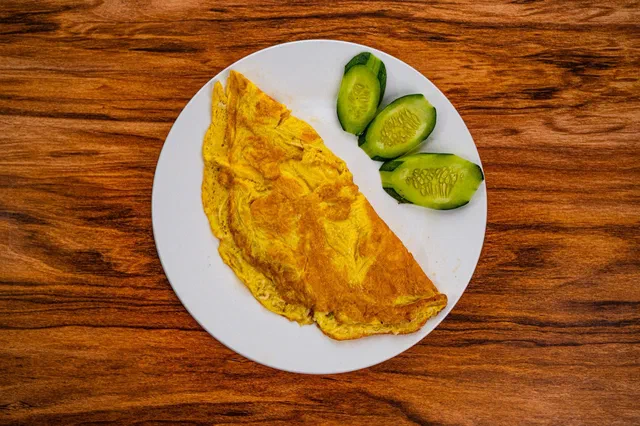 Une omelette nature pliée en demi-lune sur une assiette blanche, avec trois rondelles de concombre, sur une table en bois