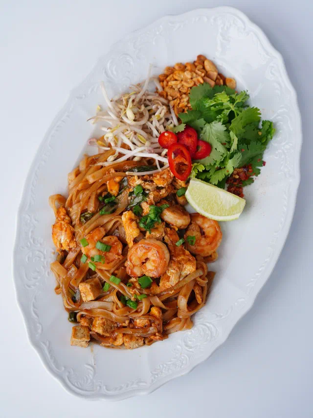 Un pad thaï aux crevettes sur un plat ovale blanc, avec pousses de soja, coriandre, piment rouge et un quartier de citron vert