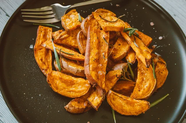 Quartiers de patate douce rôtis au four avec leur peau, parsemés de brins de romarin, dans une assiette noire avec une fourchette