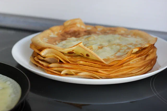 Une pile de crêpes fines et dorées sur une assiette blanche, posée sur une plaque de cuisson