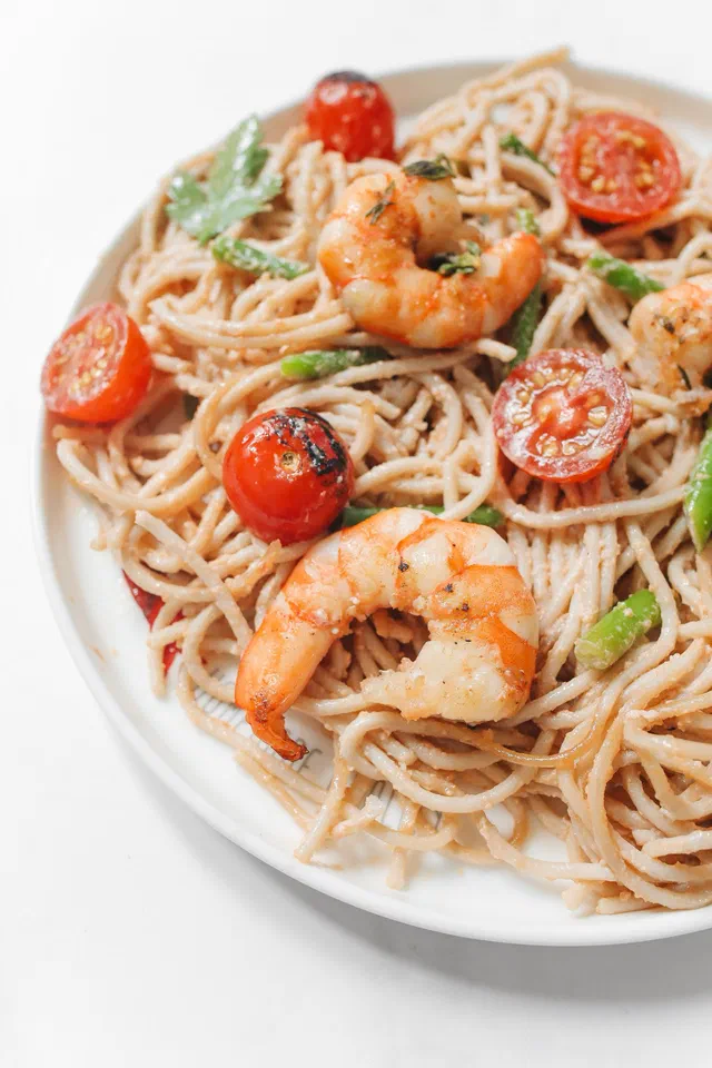 Une assiette blanche de spaghetti aux crevettes dorées, avec des tomates cerises entières et coupées, des feuilles de persil et quelques tronçons d'asperge verte