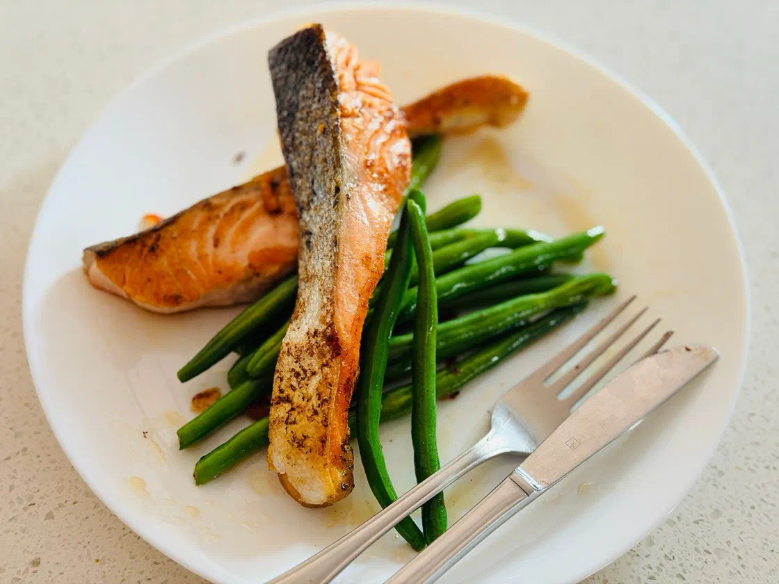 Deux morceaux de saumon poêlé à la peau grillée posés sur des haricots verts, dans une assiette blanche avec une fourchette et un couteau