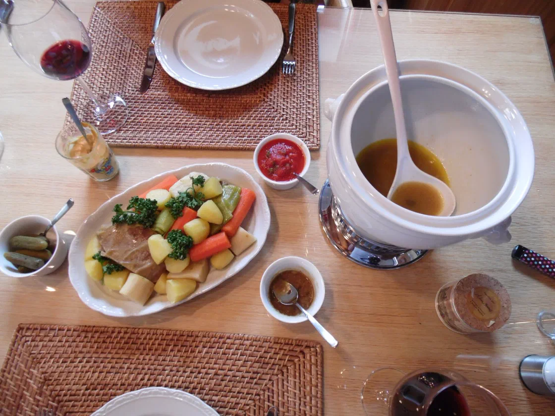 Plat de pot-au-feu avec un morceau de bœuf, carottes, navets, pommes de terre et poireaux, à côté d'une soupière de bouillon, de cornichons, de moutarde et de sauce tomate