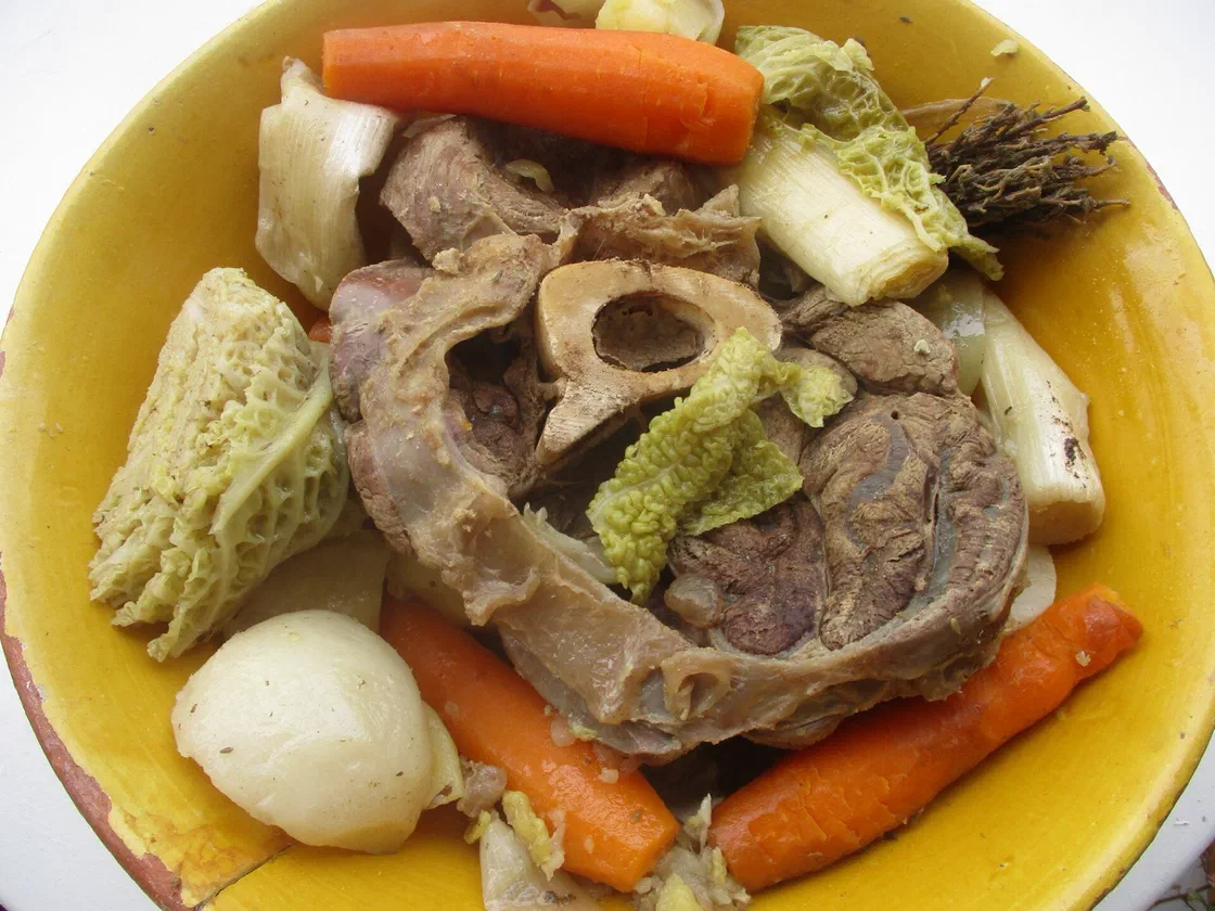 Un pot-au-feu servi dans un plat jaune : jarret de bœuf avec son os, carottes, poireaux, navet et feuilles de chou vert