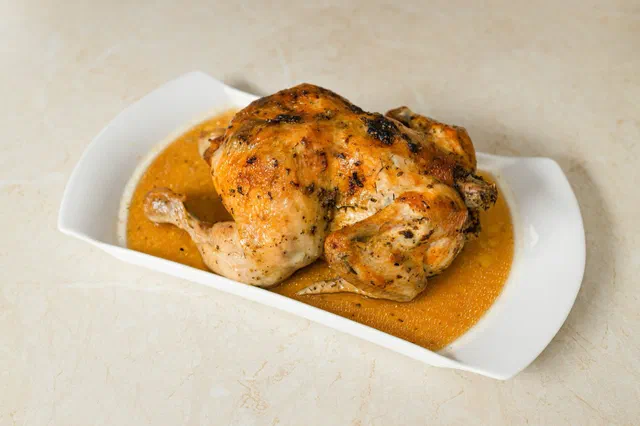 Un poulet rôti entier et doré, saupoudré d'épices, posé dans son jus sur un plat blanc allongé