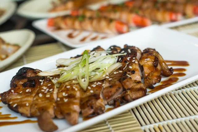 Poulet teriyaki grillé et laqué, parsemé de sésame et de fines lanières de ciboule, sur un plat blanc rectangulaire