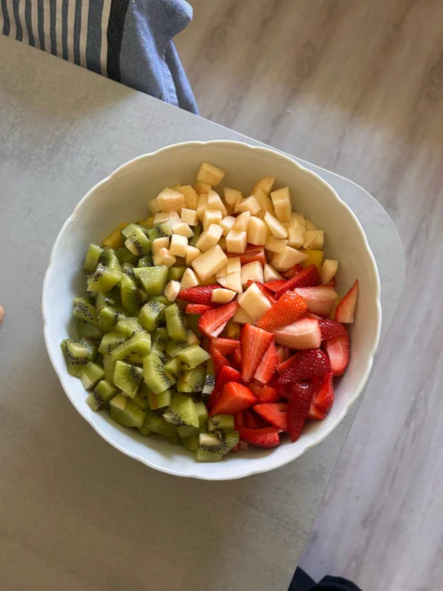 Un saladier blanc rempli de dés de kiwi, de pomme et de fraises, posé sur une table en bois clair
