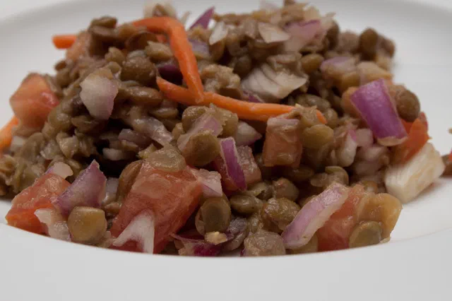 Une salade de lentilles vertes avec des dés de tomate, des lamelles d'oignon rouge et des fils de carotte dans une assiette creuse blanche