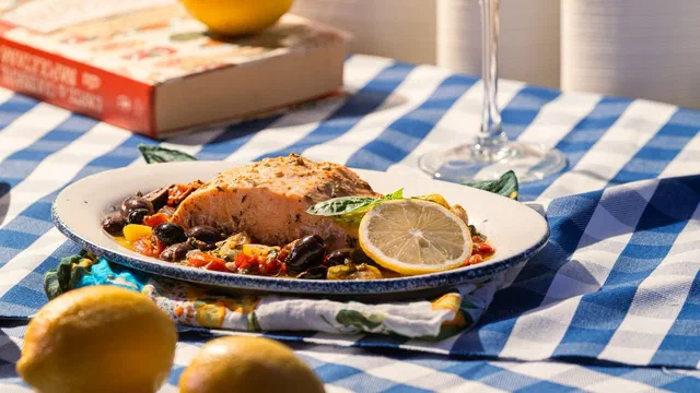 Un pavé de saumon cuit au four sur un lit de tomates confites et d'olives noires, avec une rondelle de citron et du basilic, dans une assiette émaillée posée sur une nappe à carreaux bleus