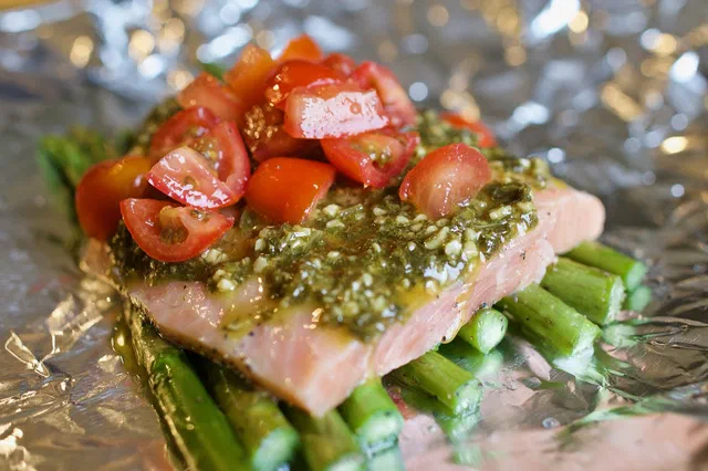 Pavé de saumon nappé de pesto et de tomates cerises en dés, posé sur des asperges vertes dans une feuille d'aluminium ouverte