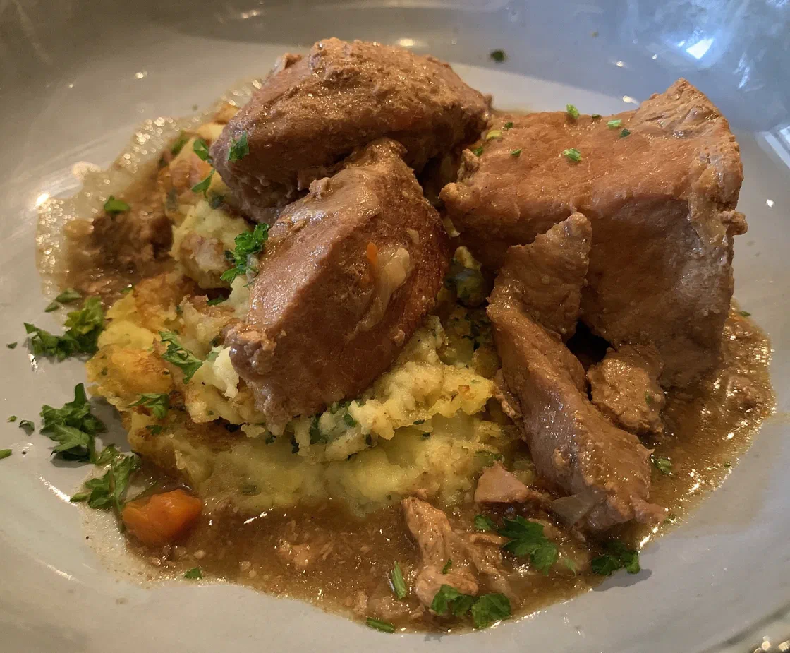 Gros morceaux de sauté de porc en sauce brune posés sur une écrasée de pommes de terre, avec du persil haché et un dé de carotte