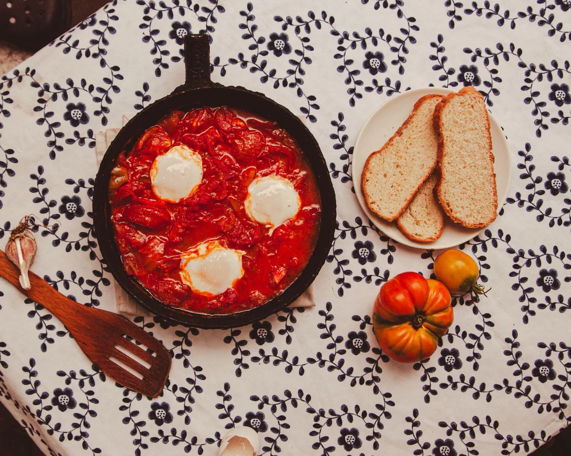 Une poêle en fonte de shakshuka vue du dessus, avec trois œufs pochés dans une sauce tomate rouge, à côté de tranches de pain