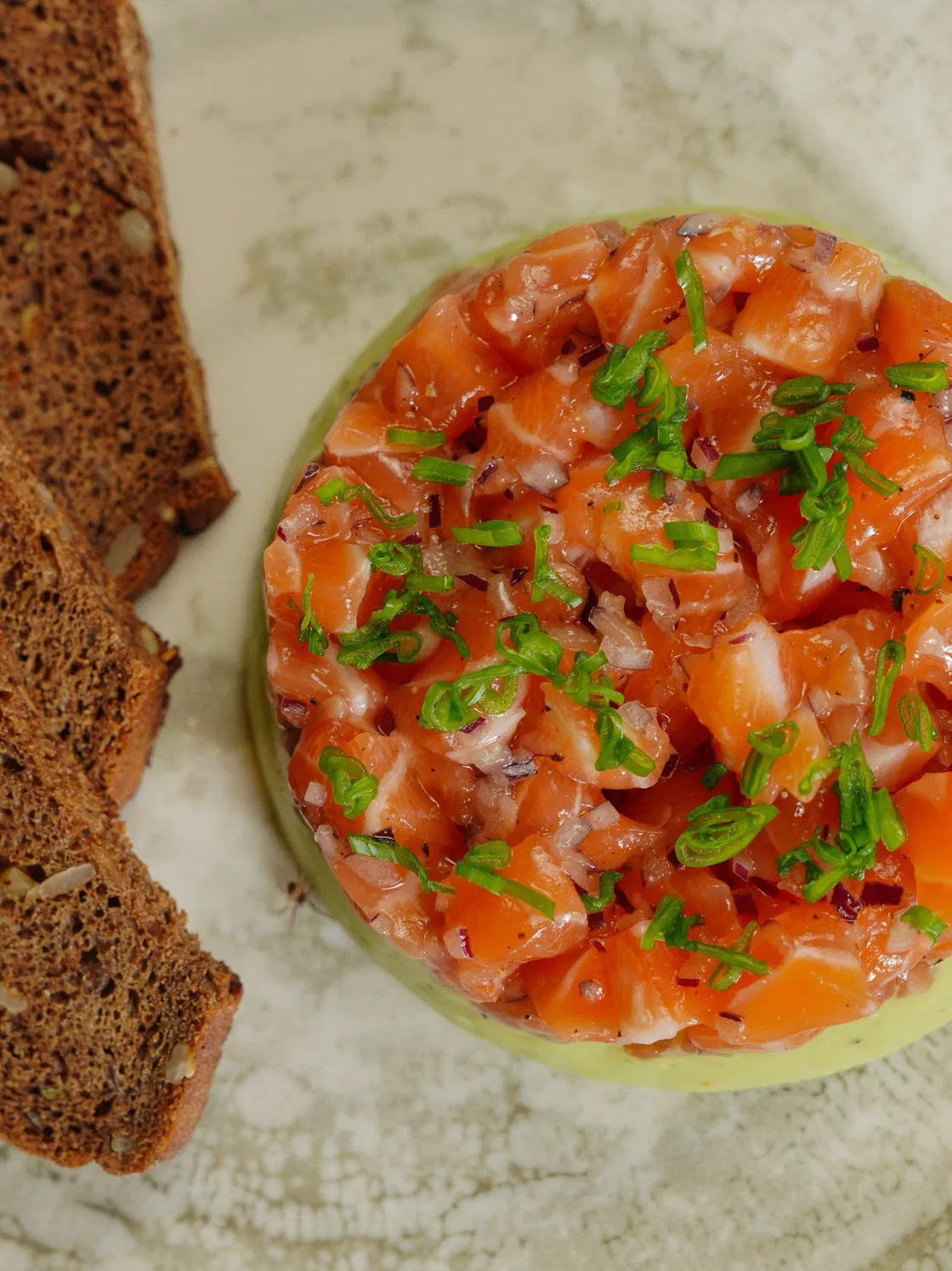 Tartare de saumon en petits dés parsemé de ciboulette ciselée, dressé en cercle sur un disque vert pâle, à côté de tranches de pain de seigle