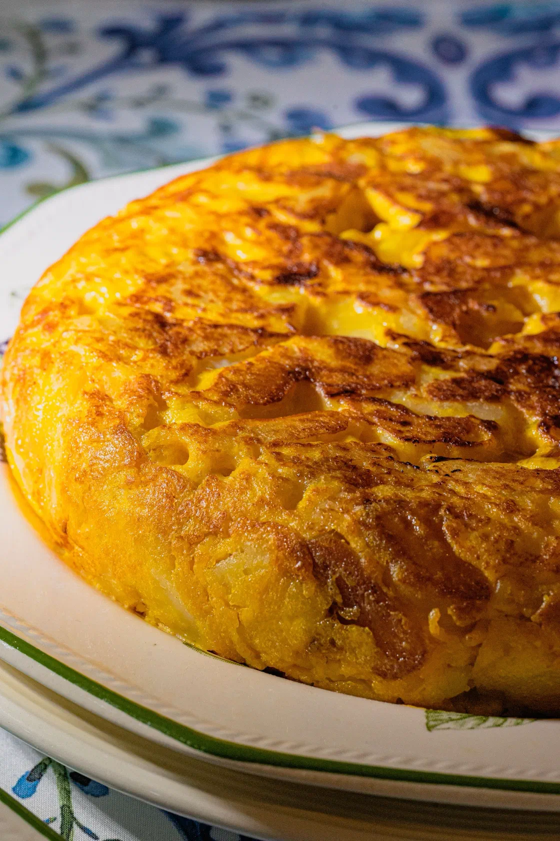 Une tortilla espagnole épaisse et dorée, aux pommes de terre bien visibles sur la tranche, posée sur une assiette blanche à liseré vert