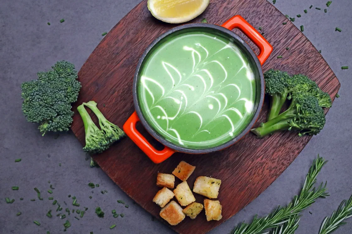 Un velouté de brocoli vert décoré de crème dans une petite cocotte rouge, entouré de bouquets de brocoli et de croûtons sur une planche en bois